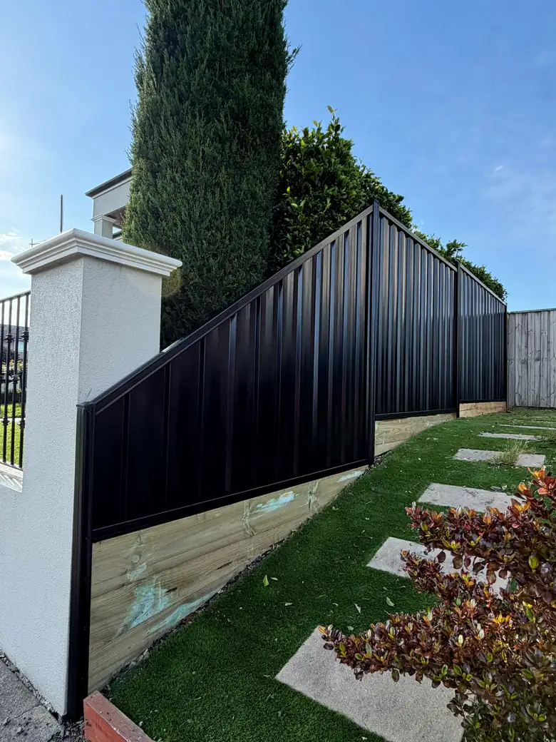 Black Colorbond fence with a timber board along the bottom, and an angled top panel dropping down to a rendered pillar