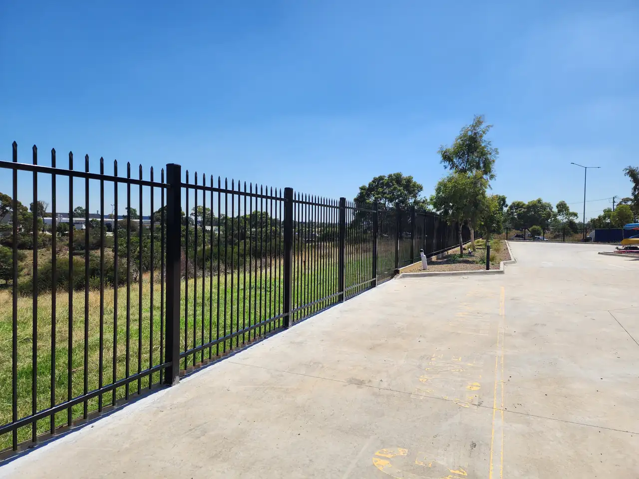 Black steel spear-top fence running along the edge of a wide concrete yard, with long grass on the other side